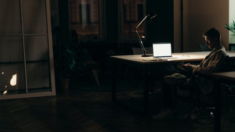 A solo founder working alone at a desk late at night, lit by a laptop screen and desk lamp