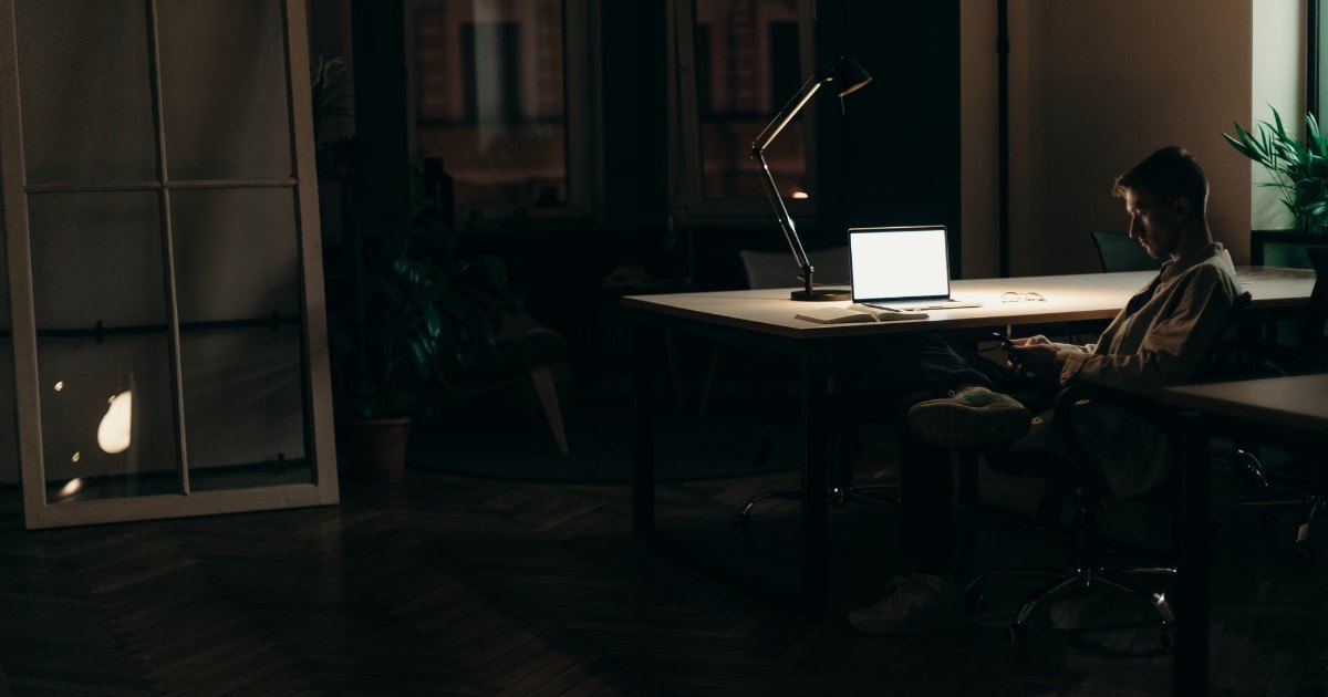 A solo founder working alone at a desk late at night, lit by a laptop screen and desk lamp